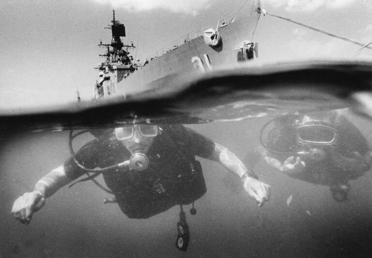 Divers swim underwater near a large ship.